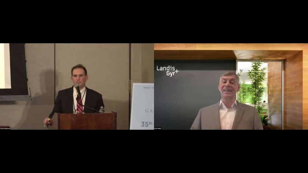 a man standing at a podium speaking and another man on a screen virtual call